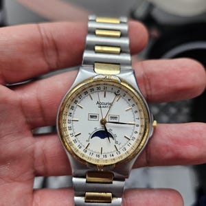 Może przedstawiać: Zegarek Accurist Quartz z białą tarczą, złotą ramką i srebrno-złotą bransoletą. Tarcza zegarka wyświetla dzień, datę i fazę księżyca. Zegarek posiada mechanizm kwarcowy.