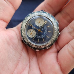 Op de afbeelding: Vintage polshorloge met een donkerblauwe wijzerplaat, drie sub-wijzerplaten en een draaiende bezel met stadsnamen. Het horloge heeft goudkleurige wijzers en markeringen, met het woord "SPORT" zichtbaar. Het horloge wordt in een hand gehouden.