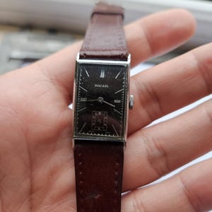 May include: A vintage rectangular wristwatch with a dark face and brown leather strap. The watch face has silver hands and the word "ROCAIL" printed on it. The watch is held in a hand.