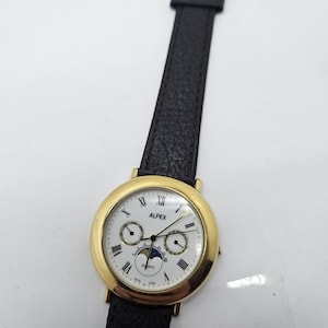 May include: Gold-tone ALFX quartz watch with a white face, Roman numeral hour markers, and a black leather strap. The watch features subdials and a moon phase indicator. The strap is approximately 23 cm long.