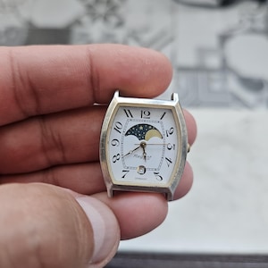 Könnte beinhalten: Ein silberfarbenes, tonnenförmiges Armbanduhrgehäuse mit weißem Zifferblatt, schwarzen arabischen Ziffern und einer Mondphasenanzeige oben. Das Zifferblatt trägt den Schriftzug Regent und Germany sowie ein kleines Datumsfenster über der Sechs.