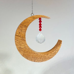 May include: A wooden crescent moon-shaped ornament with a crystal prism hanging from it. The prism is attached to a string of red beads.
