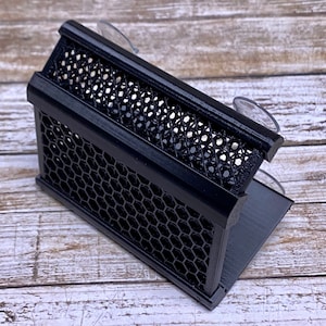 May include: A black, geometric, 3D-printed organiser with a honeycomb pattern. The organiser has a rectangular shape with a sloped base and is equipped with suction cups for mounting. The design features interconnected hexagonal shapes.