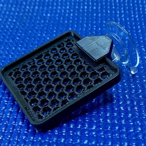 May include: A black, rectangular air vent cover with a honeycomb pattern, a black geometric handle, and a clear suction cup. The vent cover is made of plastic and sits on a blue textured surface.