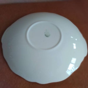 May include: White ceramic saucer with a scalloped edge. The saucer has a small, raised mark in the center.