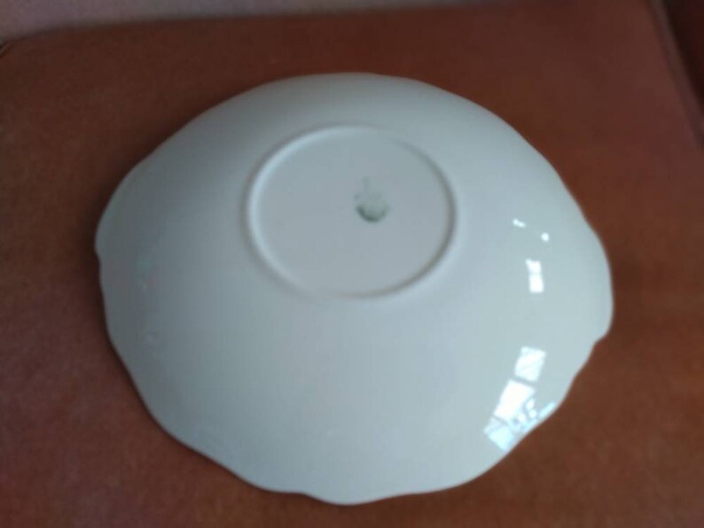 May include: White ceramic saucer with a scalloped edge. The saucer has a small, raised mark in the center.