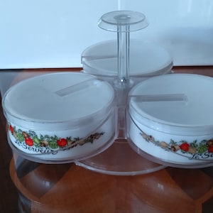 May include: A three-sectioned white serving dish set with a clear plastic rotating stand and handle. Each round bowl features a colorful vegetable print and the text Le Serveur. The clear plastic lids cover each individual white ceramic bowl.