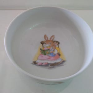 May include: White ceramic bowl featuring a cartoon illustration of two bunnies reading a book. The bunny in the rocking chair is wearing a pink dress and the smaller bunny is wearing a blue dress.