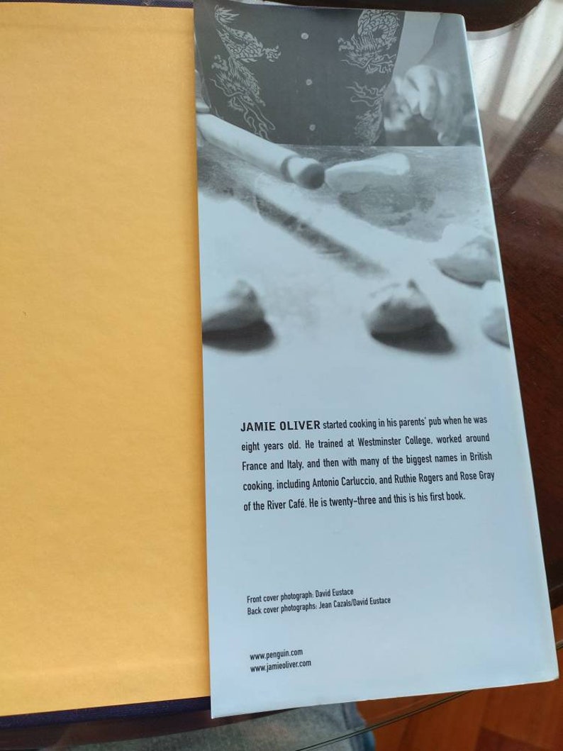 Puede incluir: Una portada de libro con una foto de un chef haciendo masa. El libro se titula "Jamie Oliver" y es su primer libro de cocina. El texto de la contraportada describe su trayectoria culinaria, incluyendo su formaci&oacute;n en el Westminster College y su trabajo con chefs famosos como Antonio Carluccio.