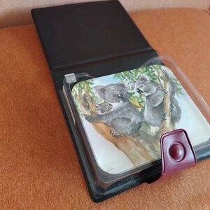 May include: A black leather photo album with a clear plastic insert featuring a watercolor painting of two koalas on a tree branch. The album has a burgundy leather closure with a circular button.