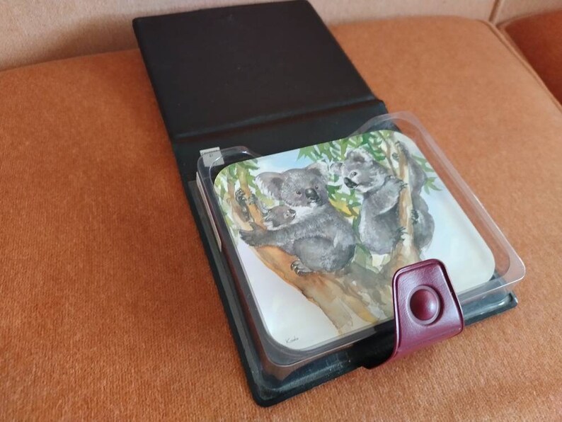 May include: A black leather photo album with a clear plastic insert featuring a watercolor painting of two koalas on a tree branch. The album has a burgundy leather closure with a circular button.