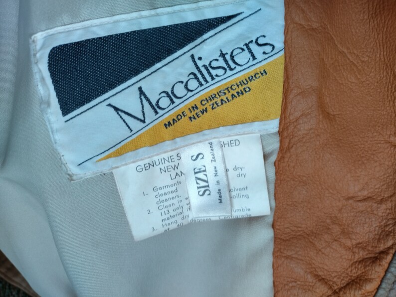 Puede incluir: Una etiqueta blanca con texto negro que dice "Macalisters Made in Christchurch New Zealand". Debajo del texto hay un tri&aacute;ngulo amarillo. Una etiqueta blanca m&aacute;s peque&ntilde;a con texto negro dice "Genuine S New LAM 1. Garments cleaned cleaners. 2. Clean in 113 only material 3. Hang dry at 40 degrees". Una tercera etiqueta blanca con texto negro dice "Size S Made in New Zealand Shed dry-dry solvent oiling Jumble Conincade".