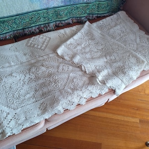 May include: Two white crocheted tablecloths with intricate floral patterns. The tablecloths are laid out on a light brown sofa.