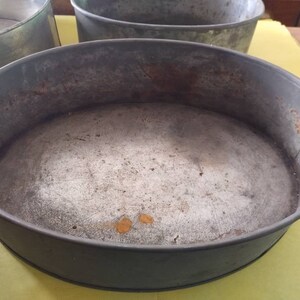 K&ouml;nnte beinhalten: Eine Auswahl an ovalen Backformen aus Metall im Vintage-Stil. Die Formen sind grau und weisen Gebrauchsspuren und Rost auf. Sie sind leer und stehen auf einer gelben Oberfl&auml;che.