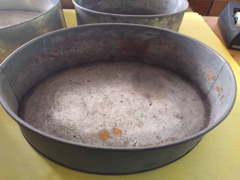 K&ouml;nnte beinhalten: Eine Auswahl an ovalen Backformen aus Metall im Vintage-Stil. Die Formen sind grau und weisen Gebrauchsspuren und Rost auf. Sie sind leer und stehen auf einer gelben Oberfl&auml;che.