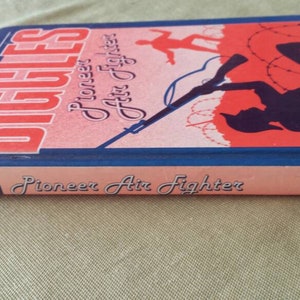 May include: A vintage book titled "Biggles Pioneer Air Fighter" by Capt. W.E. Johns. The cover features a red and blue illustration of a silhouette of a person with a rifle, barbed wire, and a plane. The book's spine is visible.