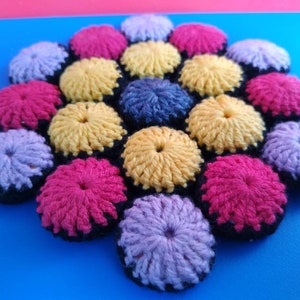 May include: A crocheted trivet with a flower design. The trivet is made of yarn in shades of pink, yellow, purple, and black.
