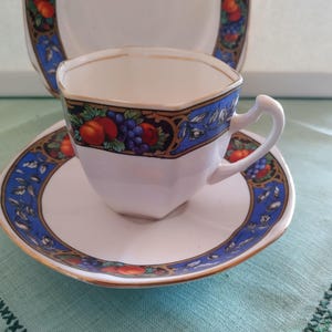 Puede incluir: Una taza de t&eacute; y un platillo blancos con un patr&oacute;n floral azul y dorado. La taza tiene una forma hexagonal y un asa. El platillo tiene un borde festoneado.