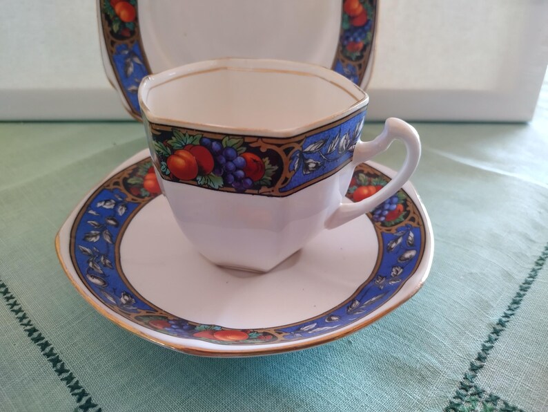 Puede incluir: Una taza de t&eacute; y un platillo blancos con un patr&oacute;n floral azul y dorado. La taza tiene una forma hexagonal y un asa. El platillo tiene un borde festoneado.