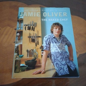 Puede incluir: Una portada de libro de cocina con Jamie Oliver, el chef desnudo, con un fondo azul y una foto de &eacute;l con una camisa de estampado azul y blanco. La portada tambi&eacute;n presenta una escena de cocina con una encimera de madera, un mortero y un pil&oacute;n, y un estante para cuchillos.