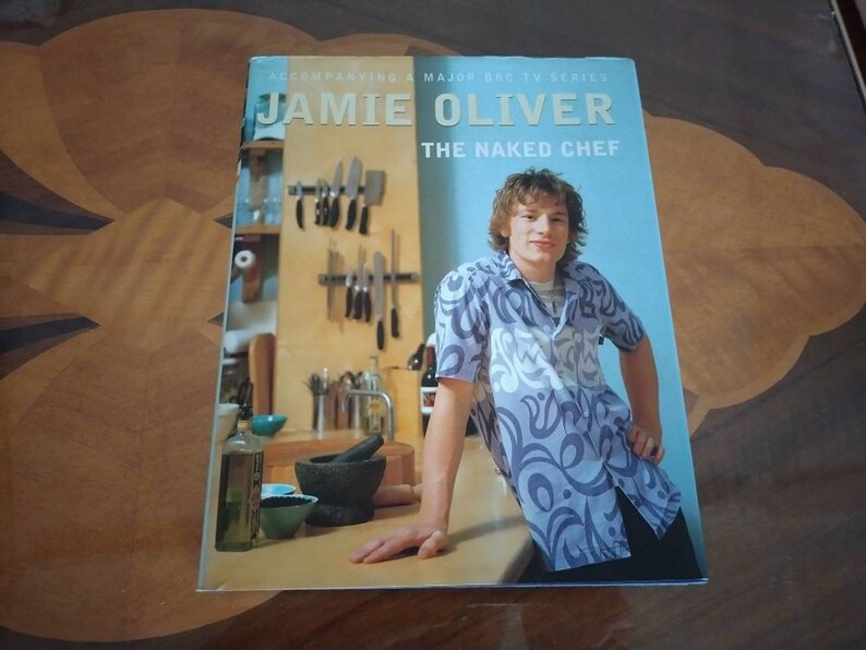 Puede incluir: Una portada de libro de cocina con Jamie Oliver, el chef desnudo, con un fondo azul y una foto de &eacute;l con una camisa de estampado azul y blanco. La portada tambi&eacute;n presenta una escena de cocina con una encimera de madera, un mortero y un pil&oacute;n, y un estante para cuchillos.