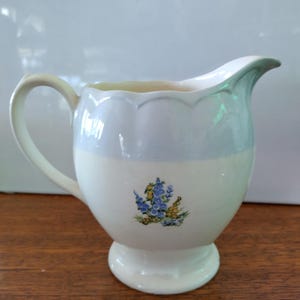 以下が含まれることがあります： 青と黄色の花柄が描かれた白い陶器のクリーマー。クリーマーは波打つ縁と足付きのベースを持っています。