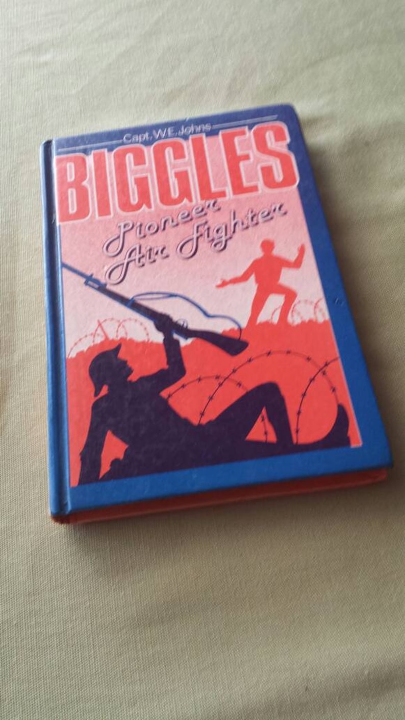 May include: A vintage hardcover book titled "Biggles Pioneer Air Fighter" by Capt. W.E. Johns. The cover features a red and blue design with a silhouette of a soldier aiming a rifle, and a running figure. The book has a blue spine.