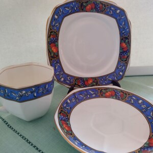 Puede incluir: Un juego de taza y platillo de t&eacute; blanco con un borde azul y dorado con un dise&ntilde;o de frutas y flores. La taza de t&eacute; tiene forma cuadrada y el platillo es redondo.