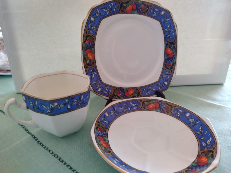 Puede incluir: Un juego de taza y platillo de t&eacute; blanco con un borde azul y dorado con un dise&ntilde;o de frutas y flores. La taza de t&eacute; tiene forma cuadrada y el platillo es redondo.