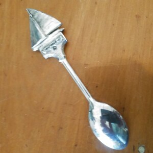 May include: A silver spoon with a sailboat design on the handle. The spoon is on a wooden surface.