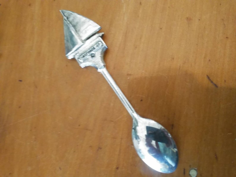 May include: A silver spoon with a sailboat design on the handle. The spoon is on a wooden surface.