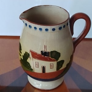 May include: A ceramic jug with a cream-coloured body, featuring a hand-painted landscape scene with a house, trees, and a red roof. The handle is brown, and the rim is decorated with blue dots. The jug has a spout for easy pouring.