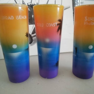 May include: Three colorful shot glasses with sunset scenes and beach-themed text. The glasses are yellow, orange, pink, blue, and purple. The text on the glasses reads "Surfers Paradise", "Gold Coast", and "Surferoad Beach".