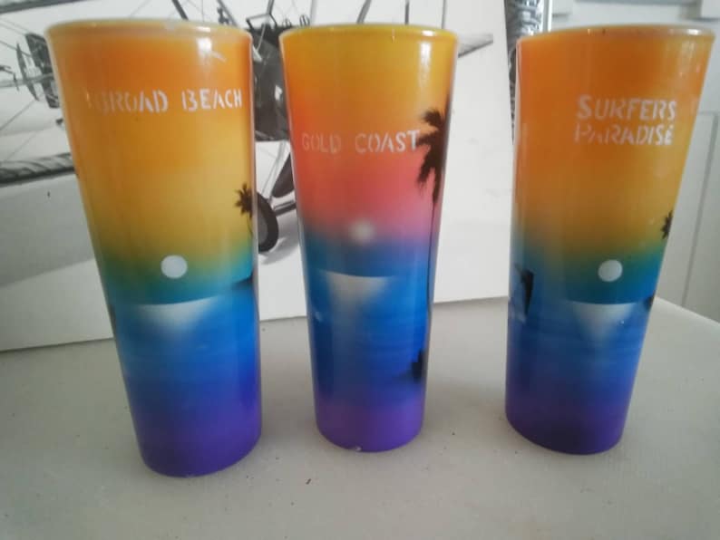 May include: Three colorful shot glasses with sunset scenes and beach-themed text. The glasses are yellow, orange, pink, blue, and purple. The text on the glasses reads "Surfers Paradise", "Gold Coast", and "Surferoad Beach".