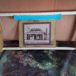 May include: A wooden embroidery frame holding a piece of tan fabric with a brown cross-stitch pattern of a pioneer family in front of a house. The fabric is attached to the frame with a bright blue strip of fabric and features the text SEMCO 5006.