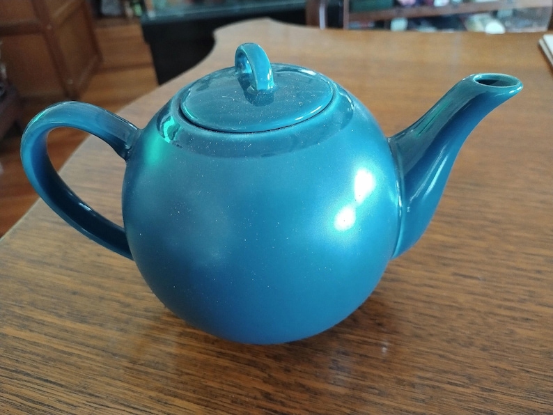 Puede incluir: Una tetera de cer&aacute;mica azul turquesa con cuerpo redondo y tapa. La tetera tiene un asa y un pico.