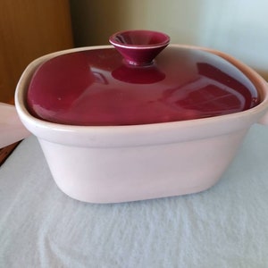 May include: A pink ceramic casserole dish with a burgundy lid. The dish has two handles on the sides and a knob on the top of the lid.