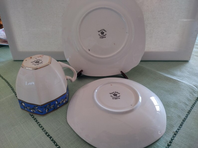 Puede incluir: Un juego de taza y platillo de t&eacute; blanco con un patr&oacute;n floral azul y dorado. La parte inferior del platillo y la taza de t&eacute; tienen impresas las palabras "Royal Albert, Crown China, England" en negro.