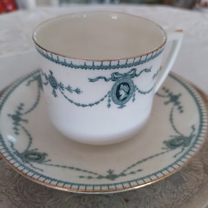 May include: A white teacup and saucer with a green and gold floral design. The saucer has a silver rim.