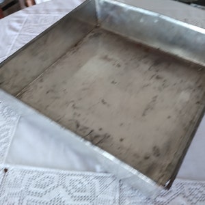May include: A rectangular, stainless steel baking tray with slightly raised edges. The tray has a worn appearance with some discolouration. The tray is on a white tablecloth with a lace pattern.