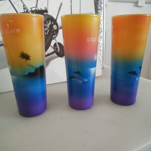 May include: Three colorful shot glasses with a sunset gradient design. The glasses feature a palm tree silhouette on the left, a dolphin silhouette on the right, and the word "Gold" on the center glass.