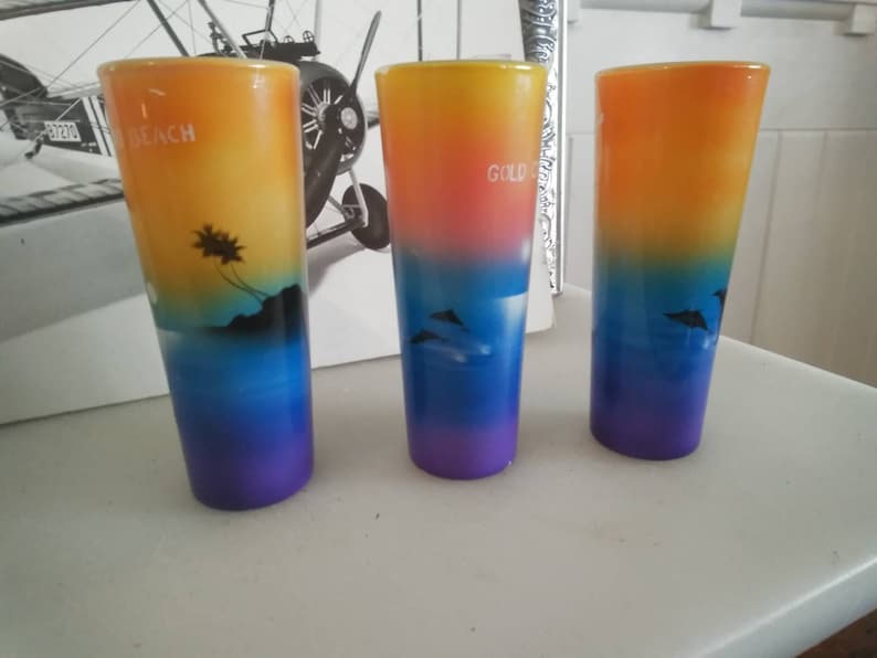 May include: Three colorful shot glasses with a sunset gradient design. The glasses feature a palm tree silhouette on the left, a dolphin silhouette on the right, and the word "Gold" on the center glass.