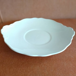 May include: A white ceramic saucer with a scalloped edge. The saucer has a slightly raised center.