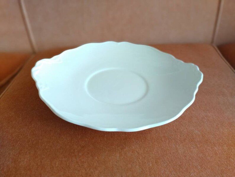 May include: A white ceramic saucer with a scalloped edge. The saucer has a slightly raised center.