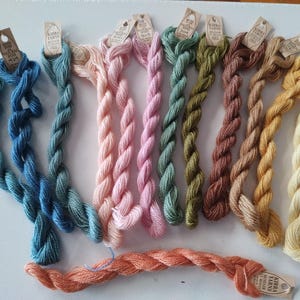May include: A collection of twelve twisted skeins of yarn in various colours including blue, pink, green, brown, yellow, and peach. Each skein features a small, light brown paper tag attached to the top, printed with the text Kirra Yarns.