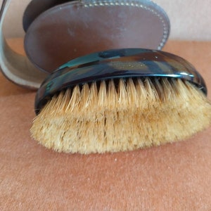 May include: A horse grooming brush with a dark, glossy handle and natural-coloured bristles. The brush is oval-shaped and appears to be made of wood and natural fibres. A brown leather strap and case are visible in the background.