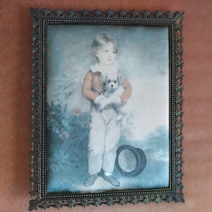 May include: A framed portrait of a young child holding a small white dog. The child is wearing a white shirt with a lace collar and a brown jacket. The portrait is set against a blue and green background.