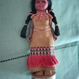 May include: A Native American doll with black hair in braids, wearing a brown suede dress with red fringe and a yellow headband. The doll has brown eyes and a painted face.