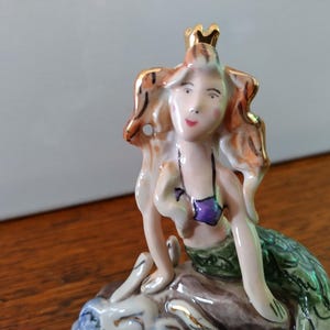以下が含まれることがあります： 金色の冠、茶色の髪、紫色の貝殻のブラジャーをつけた人魚の磁器製フィギュア。人魚は緑色の尻尾で岩の上に座っている。