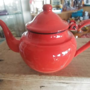 May include: A bright red enamel teapot with a black rim and a curved handle. The teapot is sitting on a wooden surface.